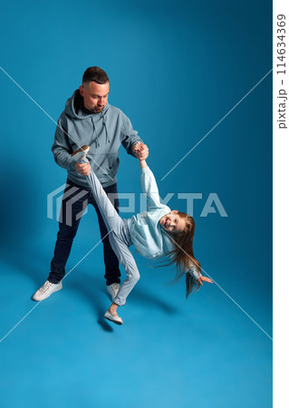 Father playing with his little daughter, holding her by hand and leg in air against blue studio background. 114634369