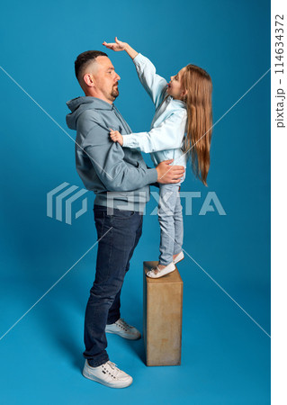 Portrait of little girl stands on stand and measures her height with her father, showing how she grew up against blue background. 114634372