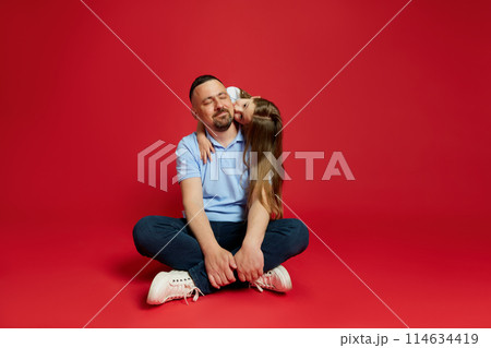 Father smiling while his beloved little daughter kissing and hugging his behind back against vibrant red background. Father smiling while his beloved little daughter kissing and hugging his behind back against vibrant red background. 114634419