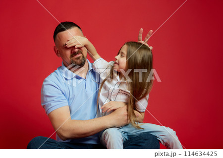 Playful moments. Charming little girl covered her father eyes while they posing against vibrant red studio background. 114634425