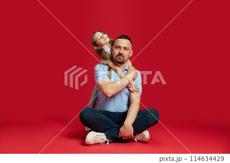 Heartwarming moments. Father sits cross-legged with his daughter embracing him from behind against vibrant red background. 114634429