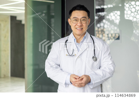 Asian mature male doctor in white coat with stethoscope smiling confidently inside a modern hospital corridor, radiating professionalism and friendliness. Asian mature male doctor in white coat with stethoscope smiling confidently inside a modern hospital corridor, radiating professionalism and friendliness. 114634869