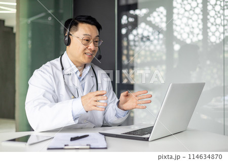 Asian mature doctor using a laptop for an online consultation, wearing a headset in a modern medical office. Asian mature doctor using a laptop for an online consultation, wearing a headset in a modern medical office. 114634870