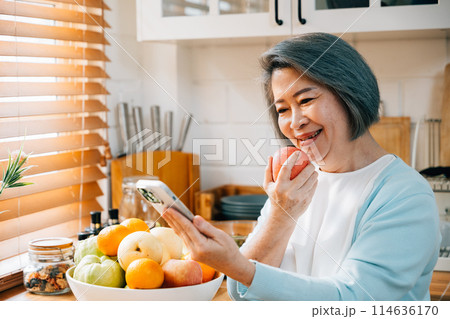 An old Asian woman, a grandmother, enjoys breakfast in the kitchen. She smiles while using her smartphone at home, eating a red apple. A portrait of modern technology and happiness. An old Asian woman, a grandmother, enjoys breakfast in the kitchen. She smiles while using her smartphone at home, eating a red apple. A portrait of modern technology and happiness. 114636170