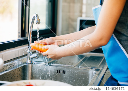 A young woman cleans and washes carrots in the kitchen highlighting organic food prep and hygiene. Illustrating washing vegetables to remove pesticides before cooking. A young woman cleans and washes carrots in the kitchen highlighting organic food prep and hygiene. Illustrating washing vegetables to remove pesticides before cooking. 114636171