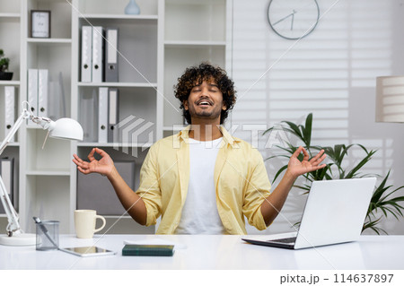 A man in a yellow shirt practicing mindfulness meditation at his desk. He is sitting in a modern office environment and appears relaxed and focused. 114637897