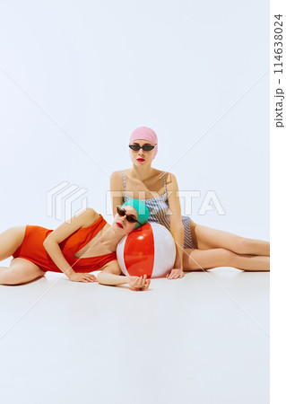 Two models in colorful swimsuits and swim caps are posing with a red and white beach ball on a white background, embodying a playful and relaxed summer vibe. Two models in colorful swimsuits and swim caps are posing with a red and white beach ball on a white background, embodying a playful and relaxed summer vibe. 114638024