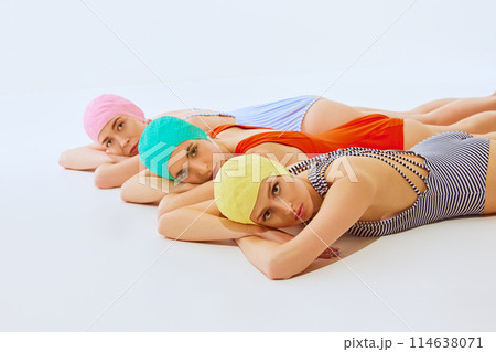 Three women, models wearing colorful swim caps and swimsuits, lying down in row on white studio background, gazing directly into camera Three women, models wearing colorful swim caps and swimsuits, lying down in row on white studio background, gazing directly into camera 114638071
