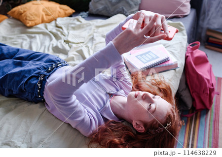 Modern teen girl wearing casual clothes lying on bed in her room taking selfies on smartphone, high angle shot Modern teen girl wearing casual clothes lying on bed in her room taking selfies on smartphone, high angle shot 114638229