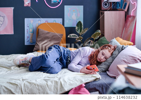 Modern Caucasian teen girl spending day indoors relaxing on bed in her bedroom watching videos in social media on smartphone 114638232