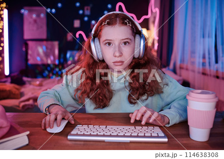 Medium POV shot of modern serious teen girl sitting at desk in her room decorated with neon lights and posters playing online video game 114638308
