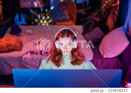 High angle view of modern teen girl wearing headphones sitting in her room surfing Internet on desktop computer and listening to music 114638317