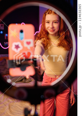 Vertical medium long shot of red-haired teen girl adjusting camera settings on her smartphone to record video for blog 114638365
