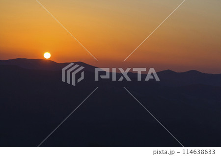Beautiful sunset in mountains. Wide panorama view of big bright white sun in dramatic orange sky over dark mountain range landscape at sunset or sunrise. Beauty of nature concept. 114638633