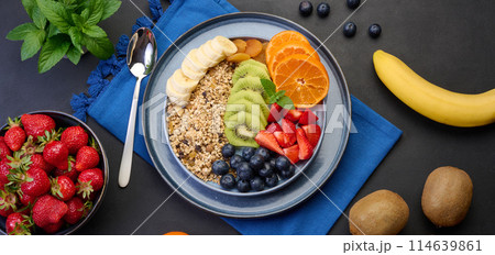Granola with strawberries, kiwi, banana and blueberries in a round plate on a black table. Healthy and tasty food Granola with strawberries, kiwi, banana and blueberries in a round plate on a black table. Healthy and tasty food 114639861