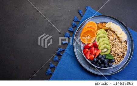 Granola with strawberries, kiwi, banana and blueberries in a round plate on a black table. Healthy and tasty food, top view Granola with strawberries, kiwi, banana and blueberries in a round plate on a black table. Healthy and tasty food, top view 114639862