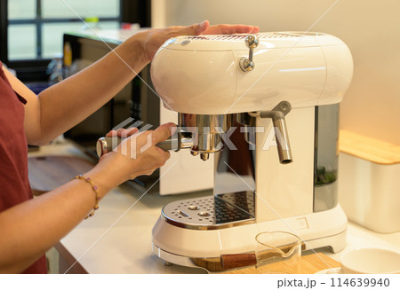 Female preparing fresh coffee while using modern coffee maker in the kitchen at home. Female preparing fresh coffee while using modern coffee maker in the kitchen at home. 114639940