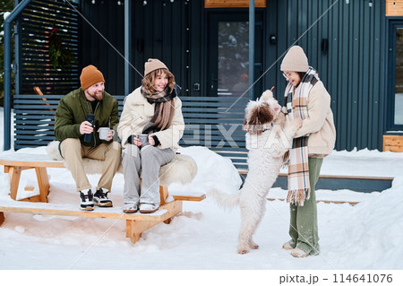 Modern Caucasian parents sitting on wooden table outdoors holding mugs with hot drink watching their daughter playing with dog on winter day 114641076