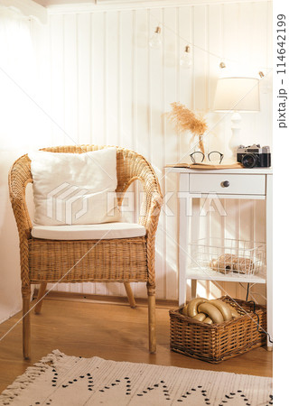 Wicker peacock chair with pillow and bedside table with book, retro camera, glasses and lamp in beige light wooden interior 114642199