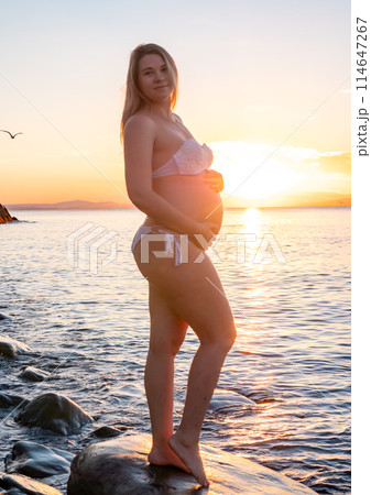 Pregnant woman in bikini posing on rocky beach at sunrise with mountain view Pregnant woman in bikini posing on rocky beach at sunrise with mountain view 114647267