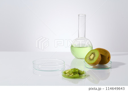 Minimalist style photo on white texture, some dishes containing fresh and chopped kiwi, a boiling flask filled by light green liquid next to an empty petri dish. Displaying space, front view 114649643