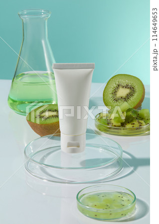 Mock up for advertising with a white color unlabeled cosmetic tube displayed on a round glass podium, an erlenmeyer flask and fresh kiwi decorated in back. Frontal shot, space for designing 114649653