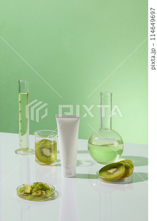 White tube without label decorated over green background with some laboratory glassware. Fresh kiwi fruit slices displayed. Empty label for cosmetic product mockup 114649697