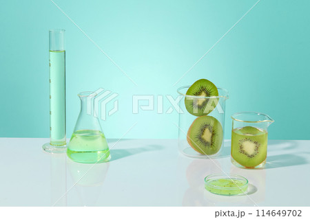 The background in light blue color features two beaker of kiwi slices and other glassware filled with liquid. Beauty product of Kiwi extract can be displayed in the blank space 114649702