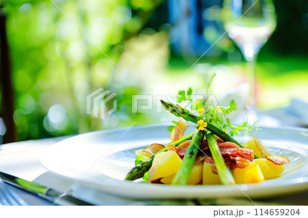 Seasonal dish of fresh asparagus on a sidewalk cafe table, an irresistible combination of asparagus, bacon and potatoes with a rich cheese sauce and a glass of white wine 114659204