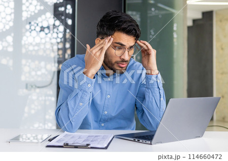A stressed businessman holding his head while working on a laptop in a modern office. Concept of stress, workload, and business challenges. A stressed businessman holding his head while working on a laptop in a modern office. Concept of stress, workload, and business challenges. 114660472