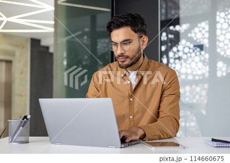 Focused professional working on a laptop in a modern, well-lit office environment. Wearing glasses and casual outfit, displaying concentration and productivity. Focused professional working on a laptop in a modern, well-lit office environment. Wearing glasses and casual outfit, displaying concentration and productivity. 114660675
