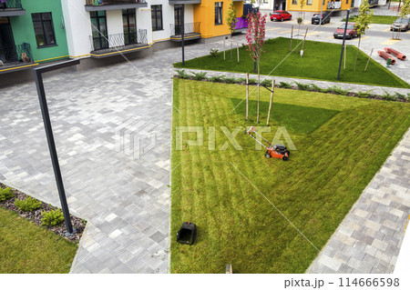 Lawn mower cutting grass on green field in yard near apartment residential building. Mowing gardener care work tool Lawn mower cutting grass on green field in yard near apartment residential building. Mowing gardener care work tool 114666598