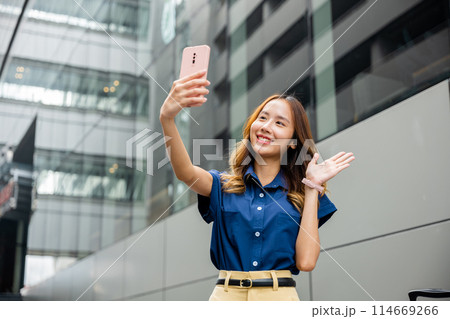 Stylish millennial woman with a bright smile, taking a selfie with her mobile phone. Youthful lady using her handheld device to capture a perfect moment in the city. 114669266