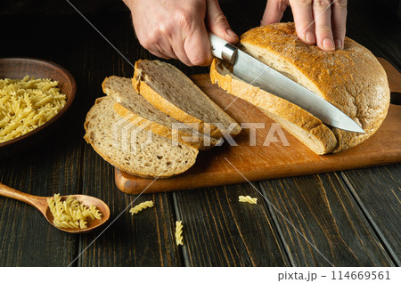 Slicing bread with a knife in the chef hand on a kitchen cutting board before dinner. Low key bakery hearty food concept. Copy space Slicing bread with a knife in the chef hand on a kitchen cutting board before dinner. Low key bakery hearty food concept. Copy space 114669561