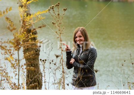 A girl stands by the lake on a warm autumn day, and points to a plant and smiles happily A girl stands by the lake on a warm autumn day, and points to a plant and smiles happily 114669641