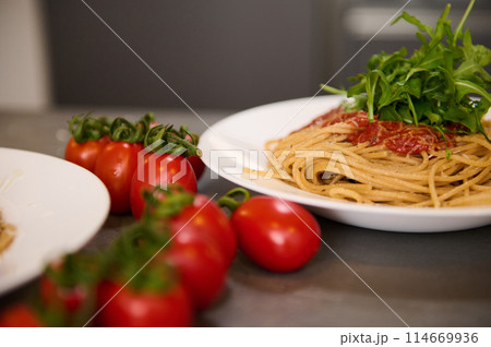 Food background. Close-up view of Italian spaghetti garnished with arugula leaves. Italian cuisine, culture and traditions. A bunch of red juicy ripe cherry tomatoes on the kitchen table Food background. Close-up view of Italian spaghetti garnished with arugula leaves. Italian cuisine, culture and traditions. A bunch of red juicy ripe cherry tomatoes on the kitchen table 114669936
