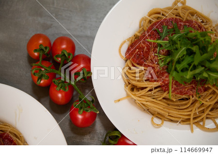 Food background with a branch of fresh ripe organic tomatoes cherry near plates of Italian pasta spaghetti a la Amatriciana with pancetta bacon and pecorino parmesan cheese. Italian cuisine. Top view. Food background with a branch of fresh ripe organic tomatoes cherry near plates of Italian pasta spaghetti a la Amatriciana with pancetta bacon and pecorino parmesan cheese. Italian cuisine. Top view. 114669947