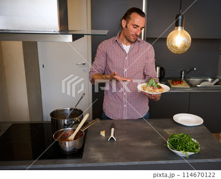 Handsome young Italian chef, man showing a white plate with Italian spaghetti capellini, standing in the home kitchen. Chef showing a served dish with Italian pasta. Italy. Food. Culture. Traditions 114670122
