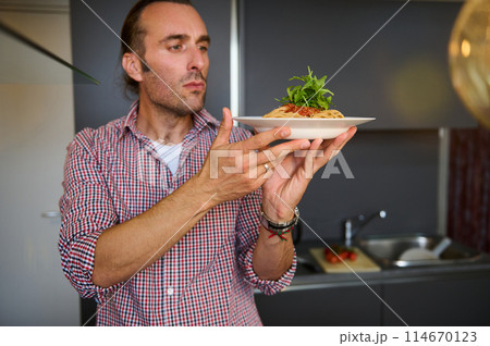 Handsome young adult man holding a white plate with Italian spaghetti capellini, standing in the home kitchen. Chef showing a served dish with Italian pasta. Italy. Food. Culture. Traditions 114670123