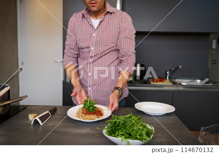 Young man cooking Italian pasta spaghetti for dinner in the home kitchen. Focus on served plate with Italian spaghetti with tomato sauce and greens arugula herbs. People, food and drink consumerism 114670132