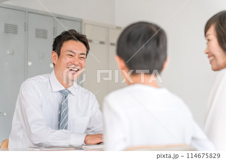 学校の教室で三者面談をする男性担任教師と男子生徒と保護者の母親 114675829