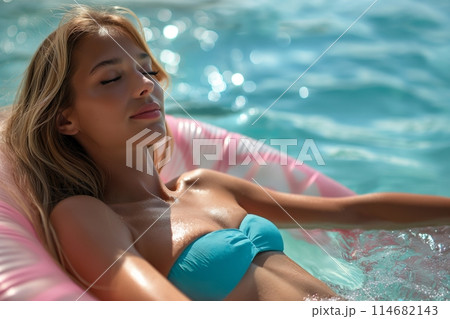 A woman in a blue bikini relaxes on a pink float in a pool, eyes closed, enjoying vacation A woman in a blue bikini relaxes on a pink float in a pool, eyes closed, enjoying vacation 114682143