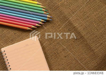 Colored pencils pastel and notebooks lay on brown crumpled burlap. 114683348