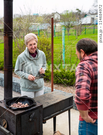 Mature russian man and woman frying meat in a cauldron on the stove outdoors, family concept 114684972