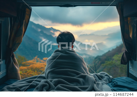 Man wrapped in a blanket enjoys a stunning mountain view from the back of his camper van at dawn 114687244
