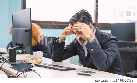 Stressed businessman sitting with head in hands at computer desk in office Stressed businessman sitting with head in hands at computer desk in office 114690715