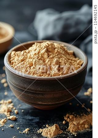 A bowl of maca powder on a textured black slate surface, with some powder sprinkled around 114692145