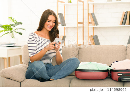 A cheerful young woman in casual clothing is seated on a beige couch, interacts with her smartphone. Open suitcases suggest she is either packing or unpacking from a trip. 114698963