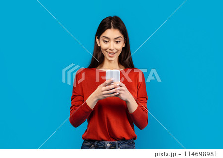 A woman wearing a red shirt is holding a cell phone in her hand, looking at the screen. She appears focused on whatever is displayed on the device, possibly texting or browsing online. 114698981