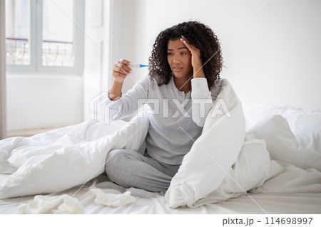 A young Hispanic woman wrapped in a white blanket is sitting up in bed with a concerned expression as she looks at a thermometer. 114698997